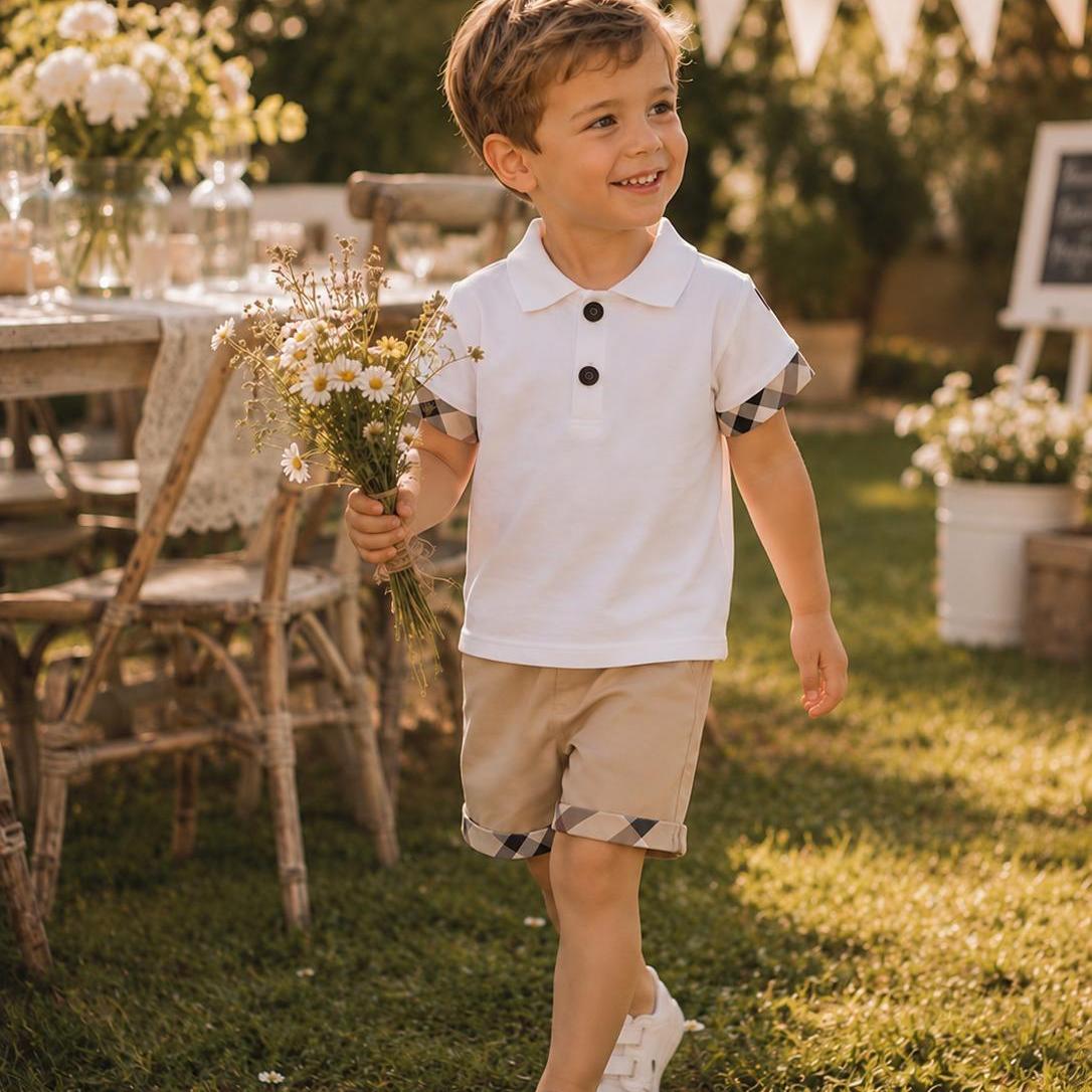A young boy smiling and walking outdoors on a grassy lawn, wearing a white short-sleeve polo shirt with patterned placket and cuffs, beige shorts, and white sneakers, while holding a small bouquet of white flowers.